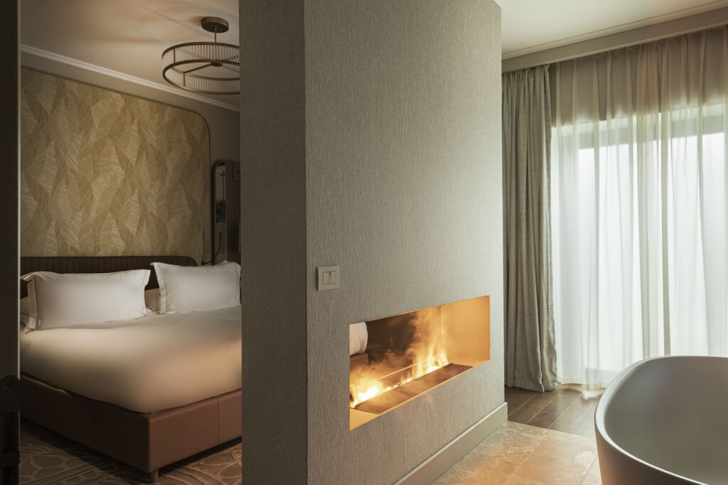 Camera da letto elegante con parete divisoria contenente un camino moderno a bioetanolo, vasca freestanding e arredi dai toni neutri.