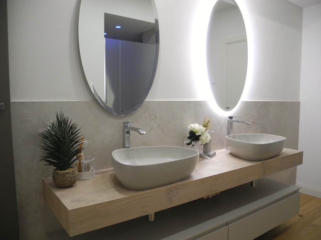Bagno moderno con doppio lavabo in ceramica su piano in legno chiaro, rubinetteria cromata e specchi ovali retroilluminati.