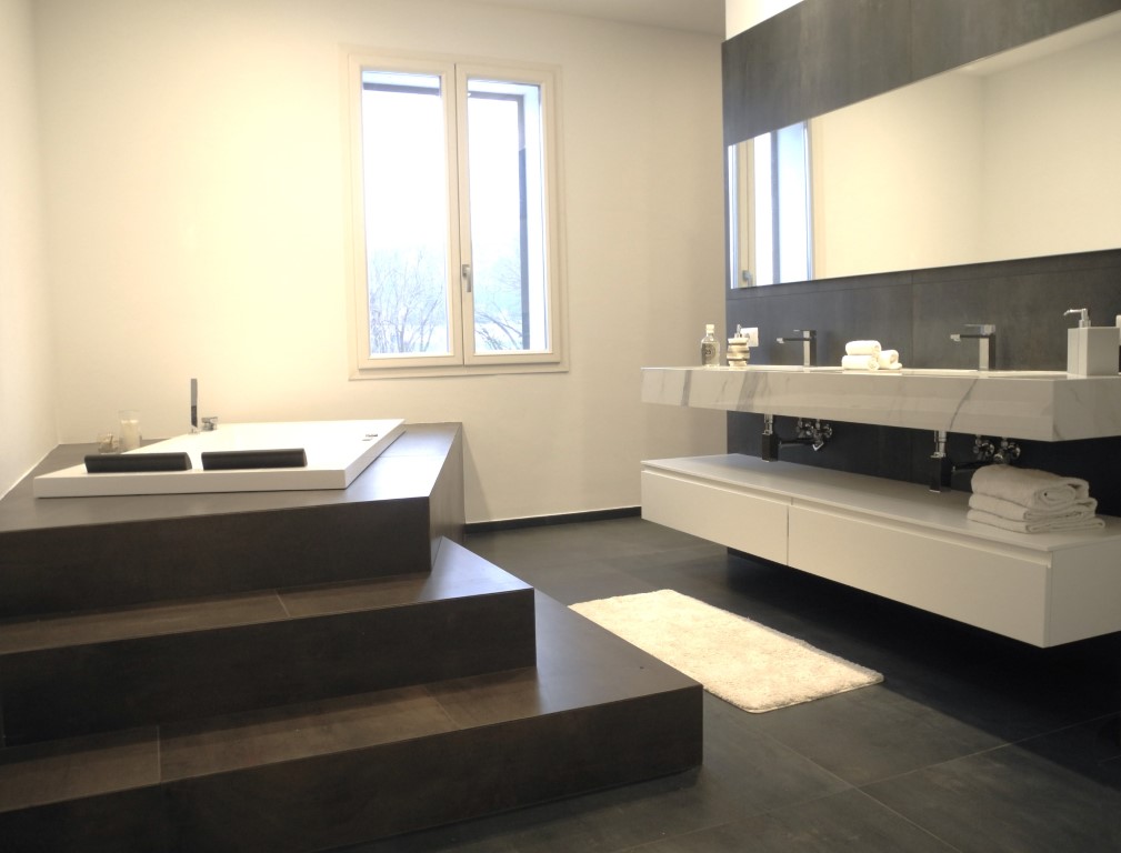 Bagno contemporaneo con vasca incassata su gradoni in gres scuro e doppio lavabo in marmo sospeso su parete grigio antracite.