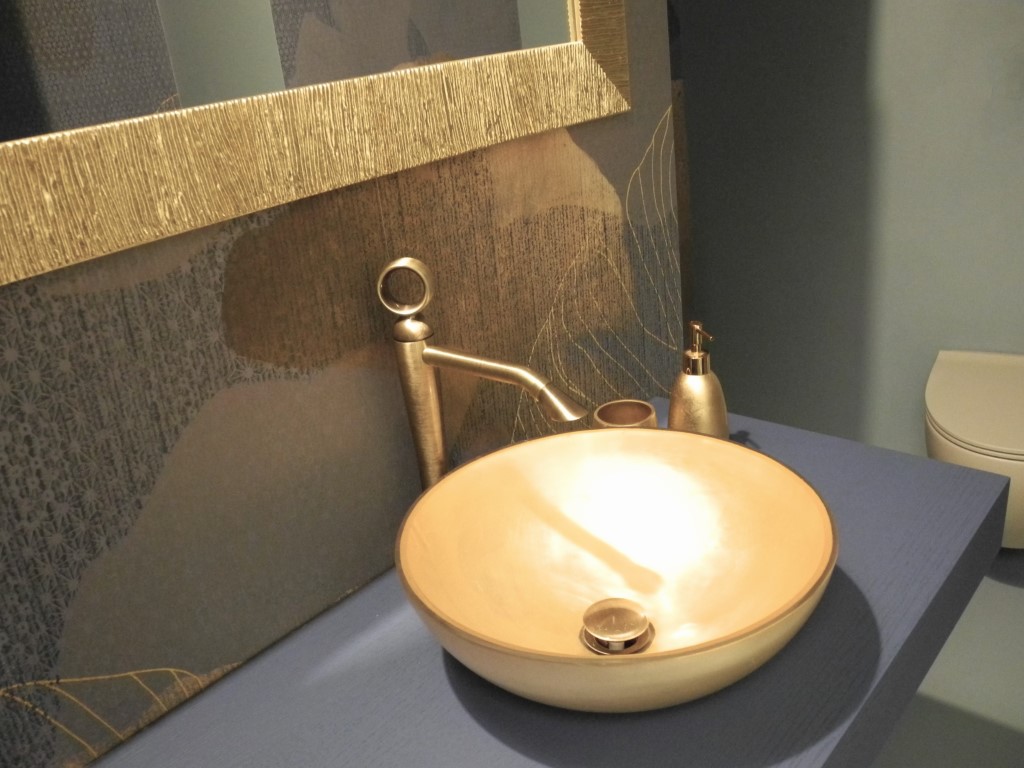 Dettaglio di lavabo dorato con rubinetteria in finitura metallica e parete decorata con rivestimento materico blu e oro.
