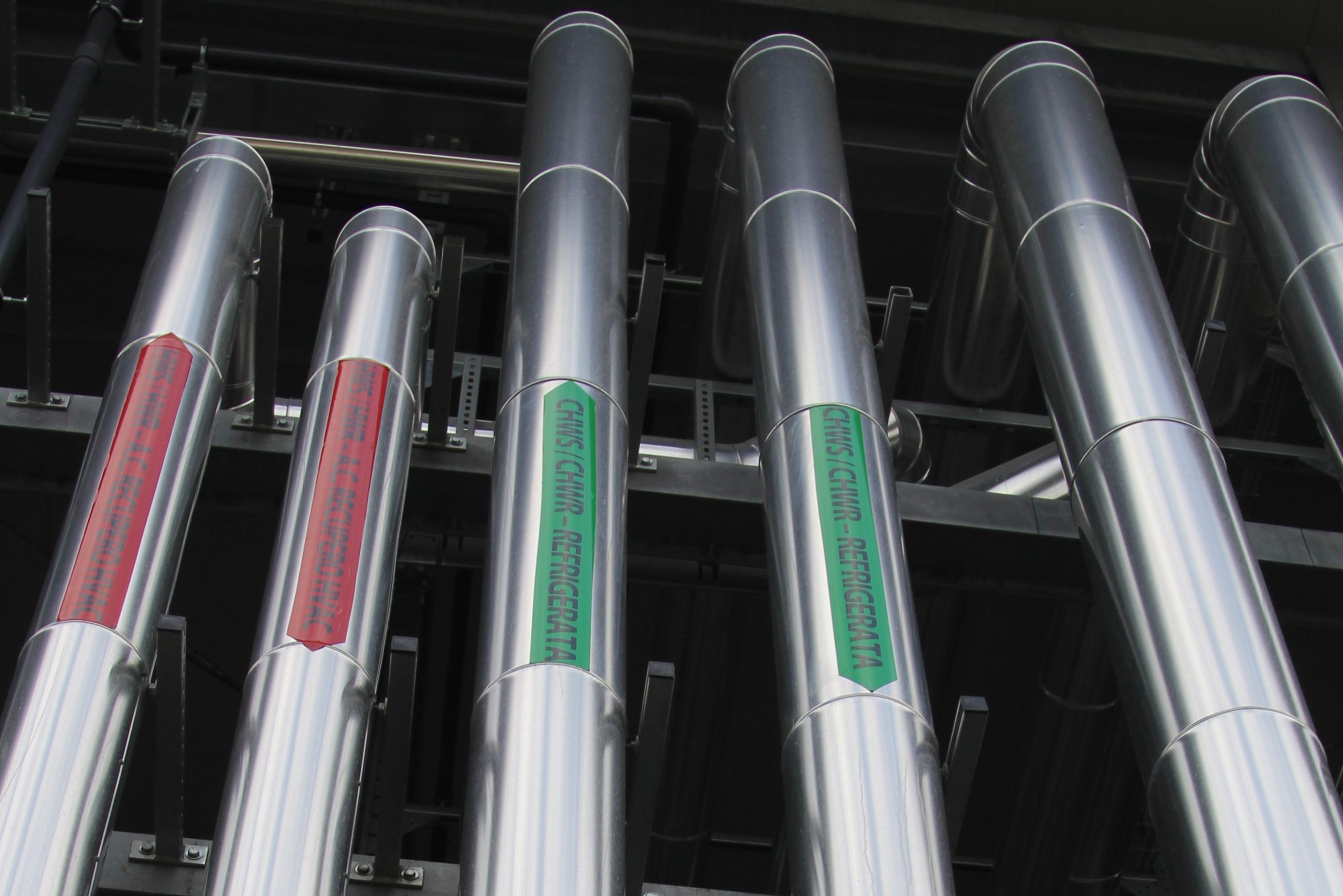 Dettaglio di tubazioni industriali in acciaio inox con etichette rosse e verdi che indicano i flussi di acqua calda e refrigerata.