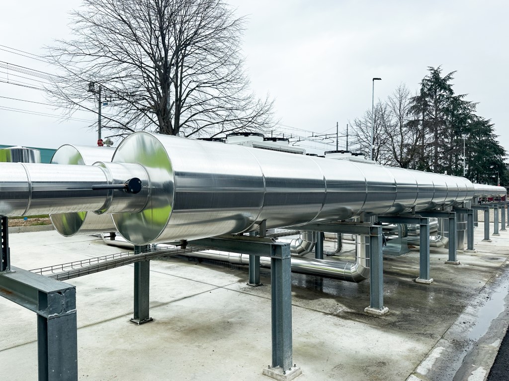 Vista esterna di un impianto industriale con tubazioni metalliche coibentate e unità di climatizzazione Aermec sullo sfondo.
