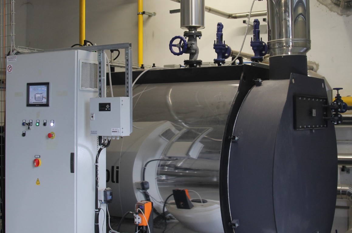 Sala impianti industriale con grande caldaia in acciaio, tubazioni isolate e quadro di controllo elettronico.